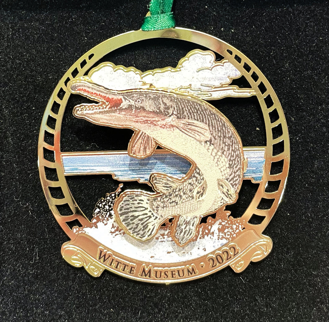 Round gold metal ornament with a brown and grey alligator gar jumping out of blue water with white clouds in the background. Ornament says 